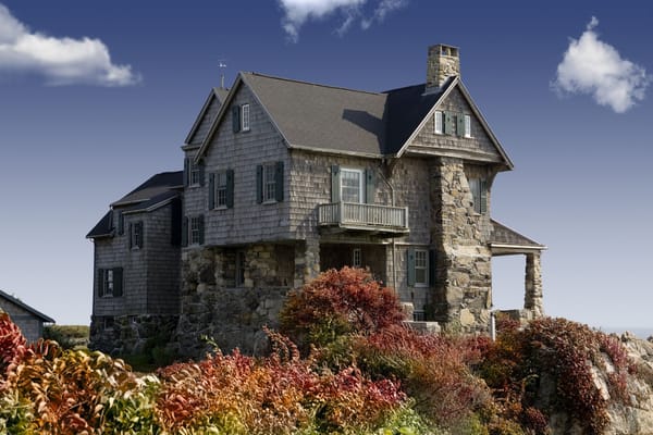 Historic stone and shingle New England home perched on rocky hillside surrounded by vibrant autumn foliage.